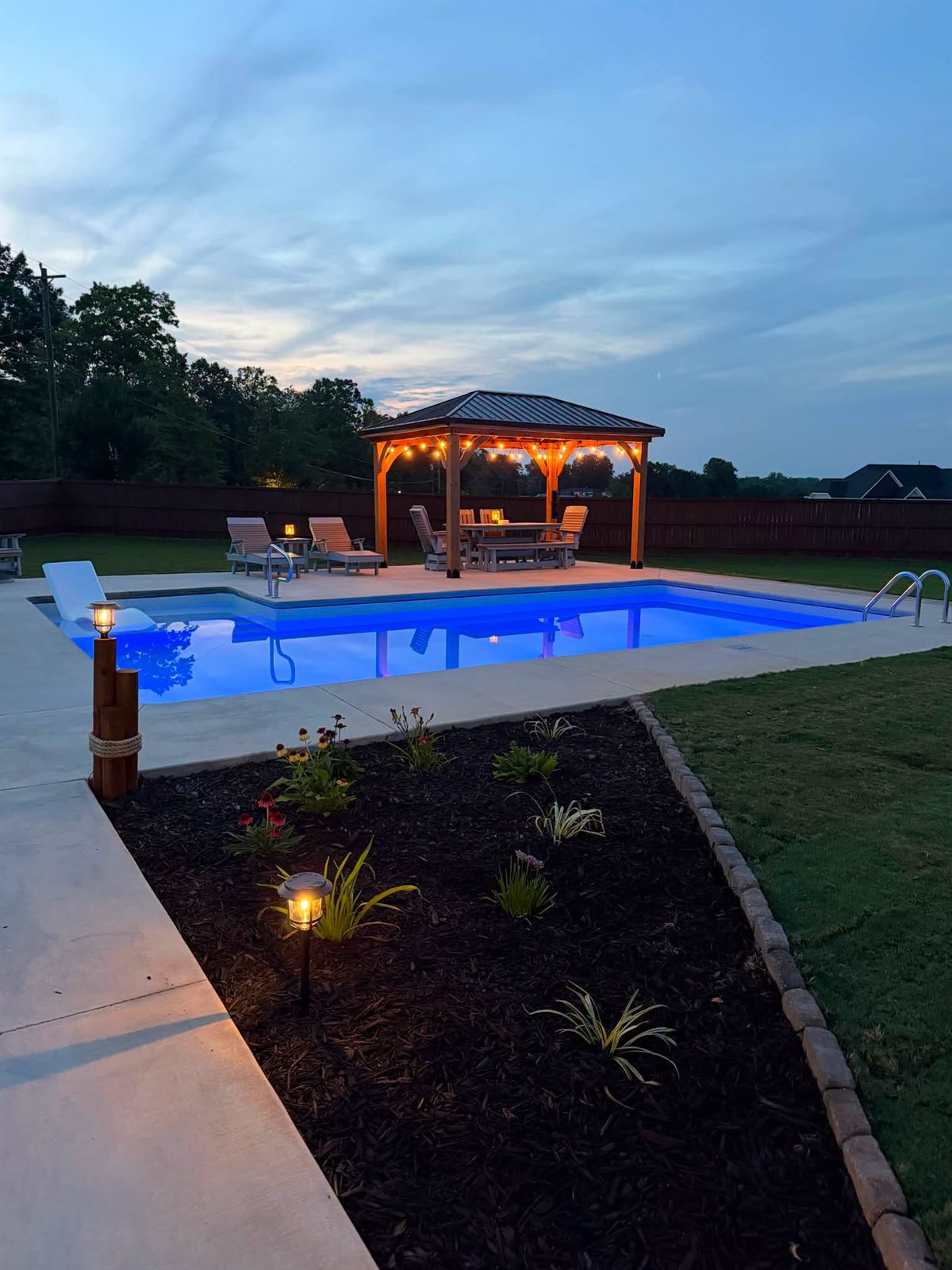 Pool lit blue at dusk with a lit gazebo, landscape lighting and a planted border