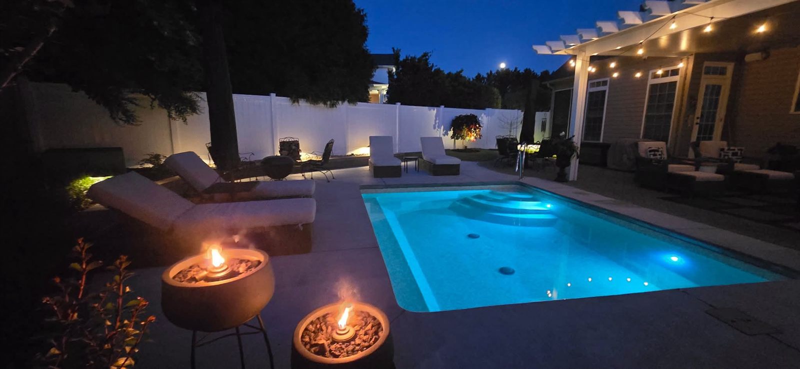 Backyard pool lit from within at night with fire bowls and string lights on the patio