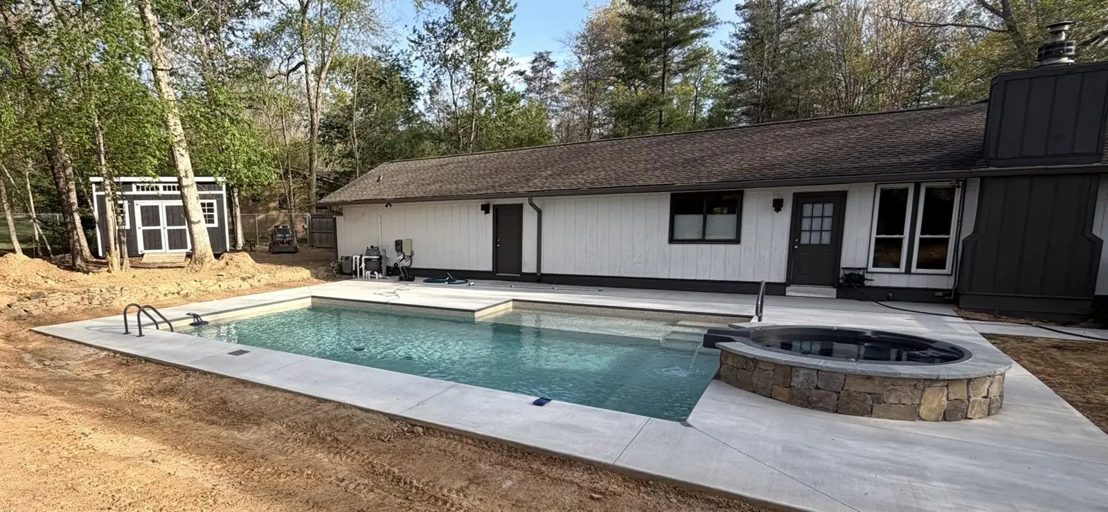Rectangular pool with a raised stacked-stone spa spilling into it, behind a white ranch home in the pines
