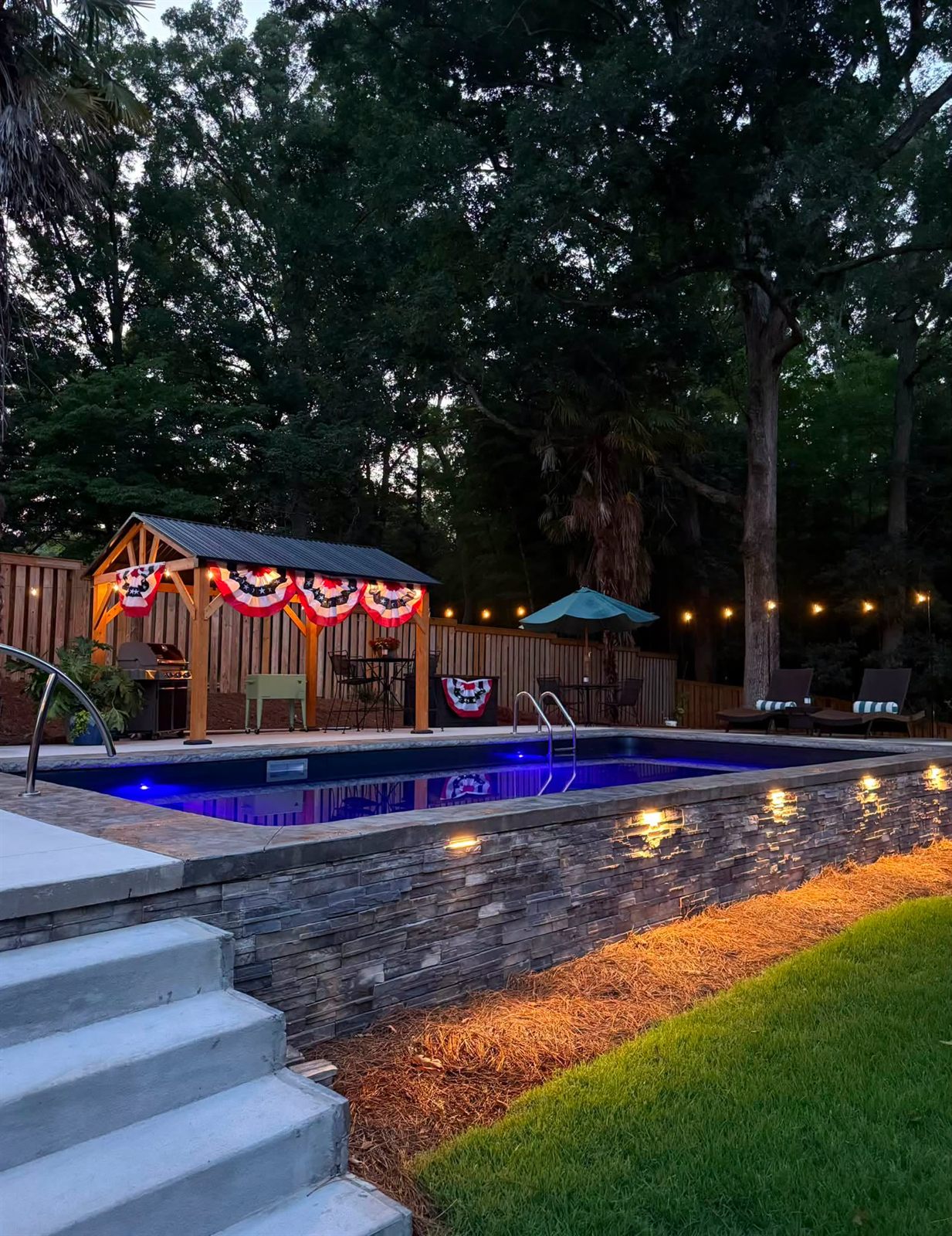 Raised pool edged by a lit stacked-stone wall with a cedar pavilion and string lights at dusk