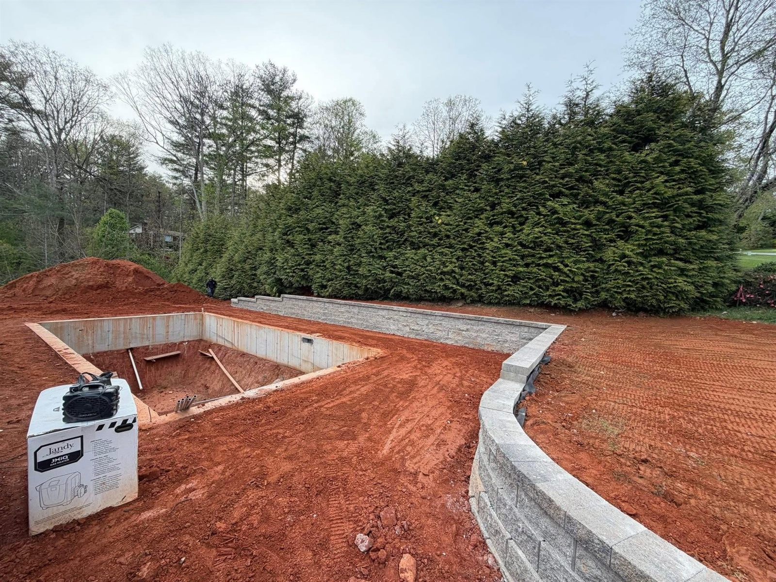 Tiered stacked-block retaining wall curving around a formed pool shell in red clay